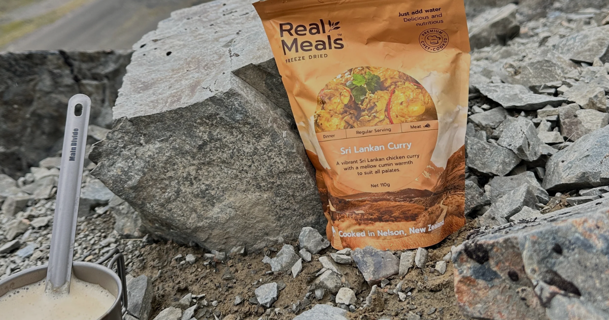 Real Meals freeze-dried Sri Lankan Curry pack propped on rocks beside a Main Divide camp mug in alpine terrain