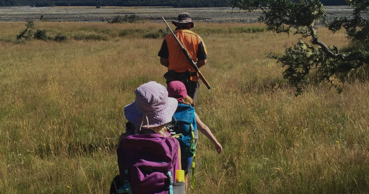 2:47 PMFamily walking through golden grasslands towards a braided river, hunter in hi-vis vest carrying a rifle leading two children with backpacks