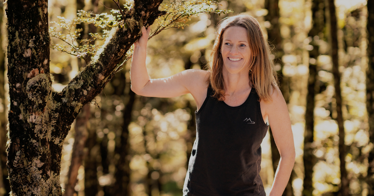 Woman wearing a Main Divide merino singlet leaning against a mossy tree in sunlit New Zealand bush
