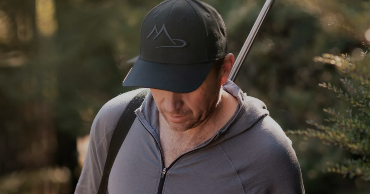 Close-up of a hunter wearing a Main Divide cap and merino zip-up hoodie carrying a rifle through bush