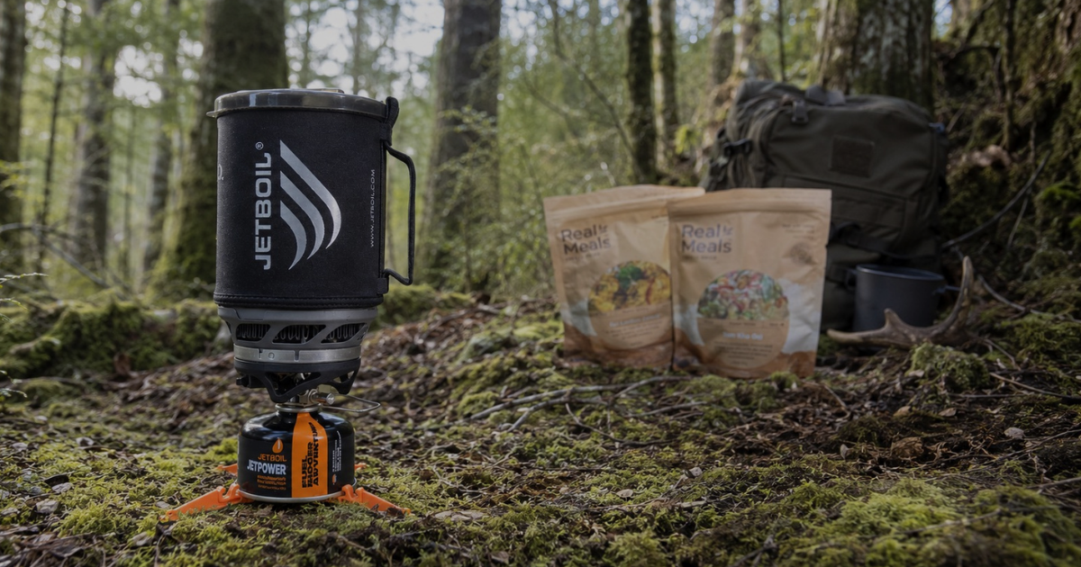 Jetboil camping stove set up on a mossy New Zealand bush floor in daylight, with native forest, a hiking pack, freeze-dried meal pouches, and camp cookware softly blurred in the background.
