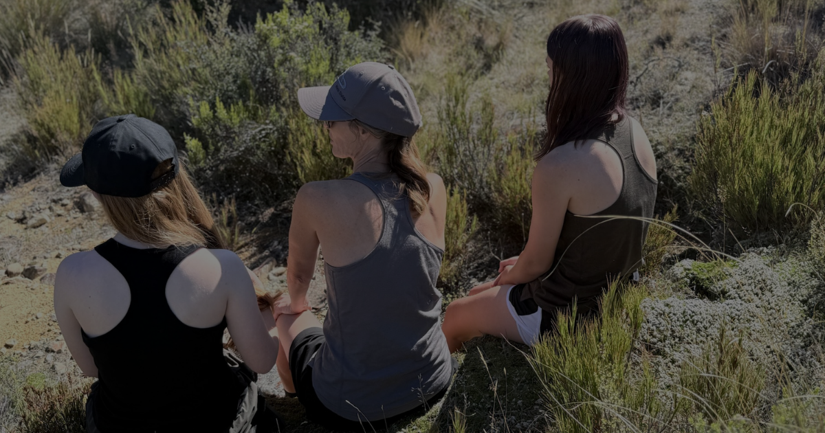 Three women in Main Divide tank tops sitting among tussock in the New Zealand high country