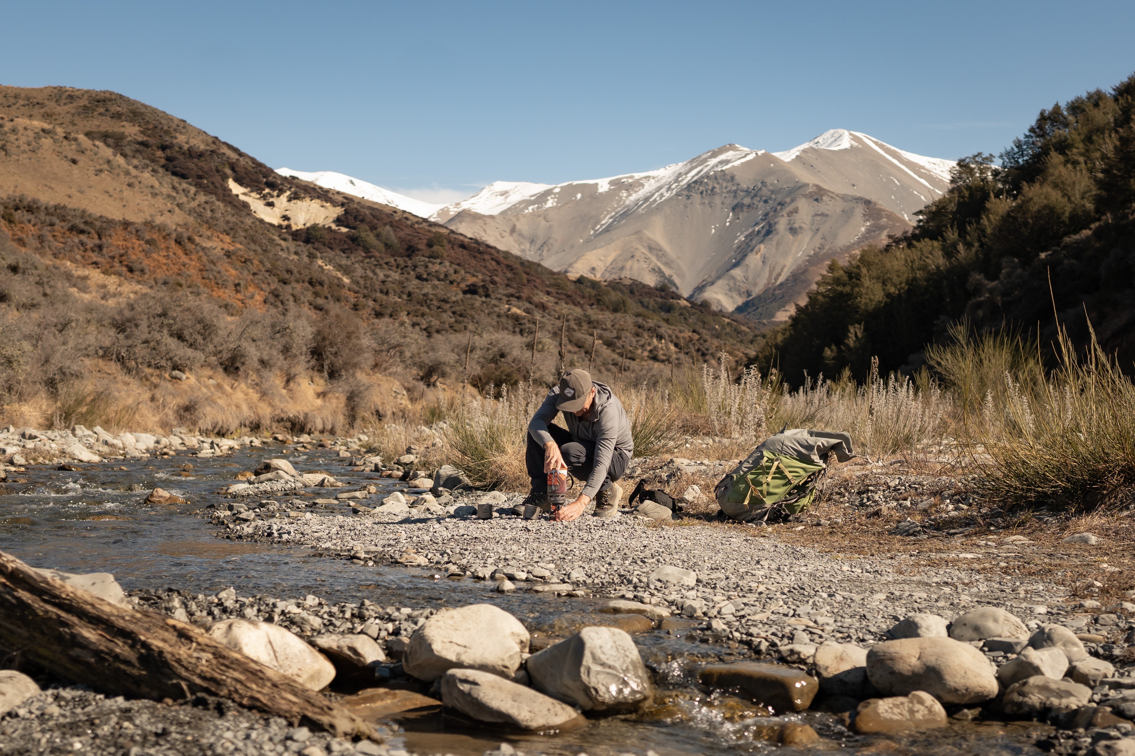 Hunter crouching beside a river in the New Zealand backcountry, with mountains in the background, taking a break during a hunt.