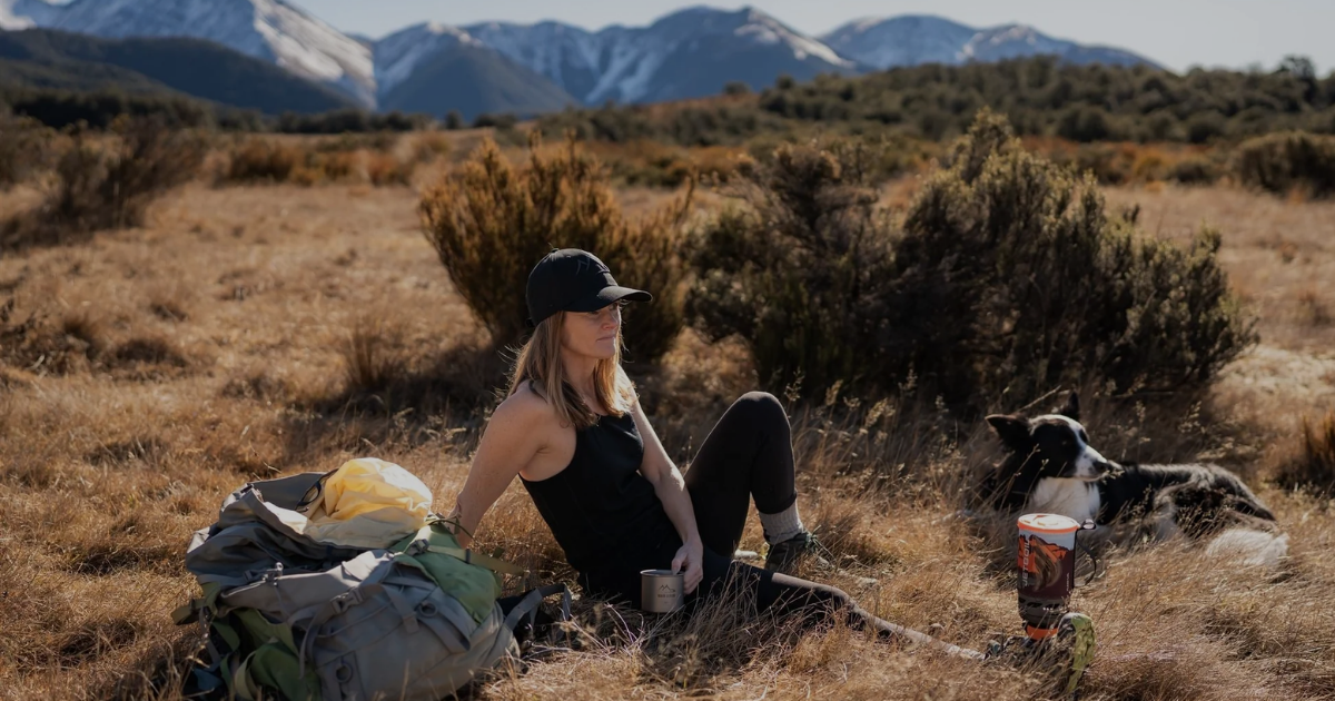 Woman resting in tussock grasslands with a backpack, camp mug and border collie, snow-capped New Zealand mountains in the background