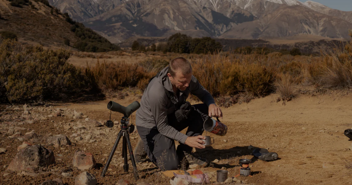 Hunter in Main Divide merino apparel making a hot drink beside a spotting scope and camp stove in dry New Zealand high country