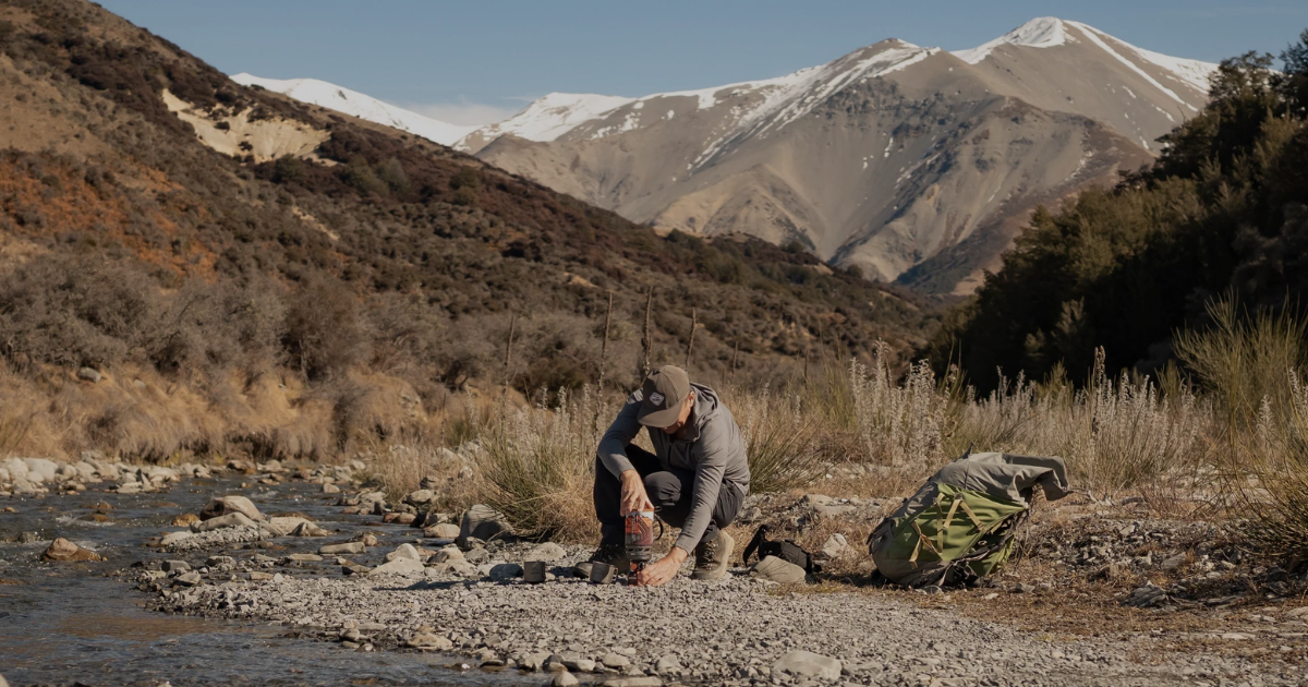 Person in Main Divide merino apparel crouching beside a rocky river in the New Zealand high country, pack on the ground and snow-capped mountains in the background