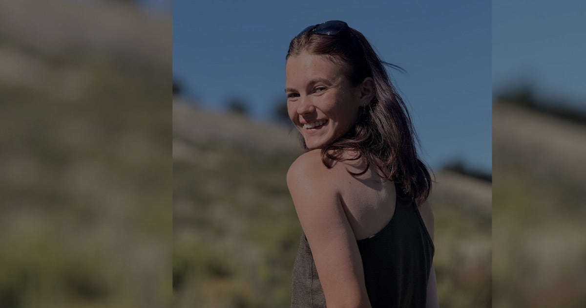 Woman wearing a black HTM 120 Nuyarn® Merino singlet, smiling over her shoulder on a sunny tussock hillside in the New Zealand high country