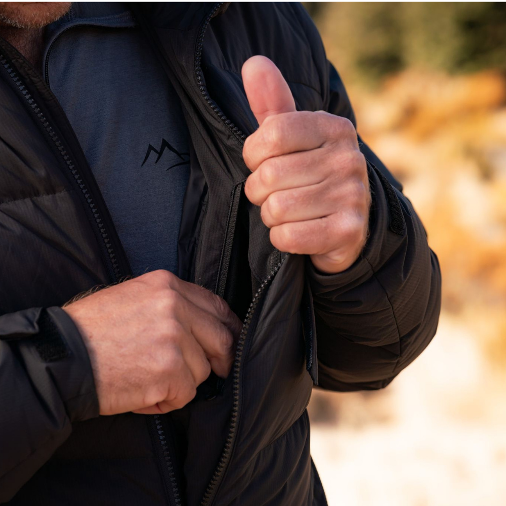 Person wearing a black jacket with a logo, giving a thumbs-up gesture outdoors.